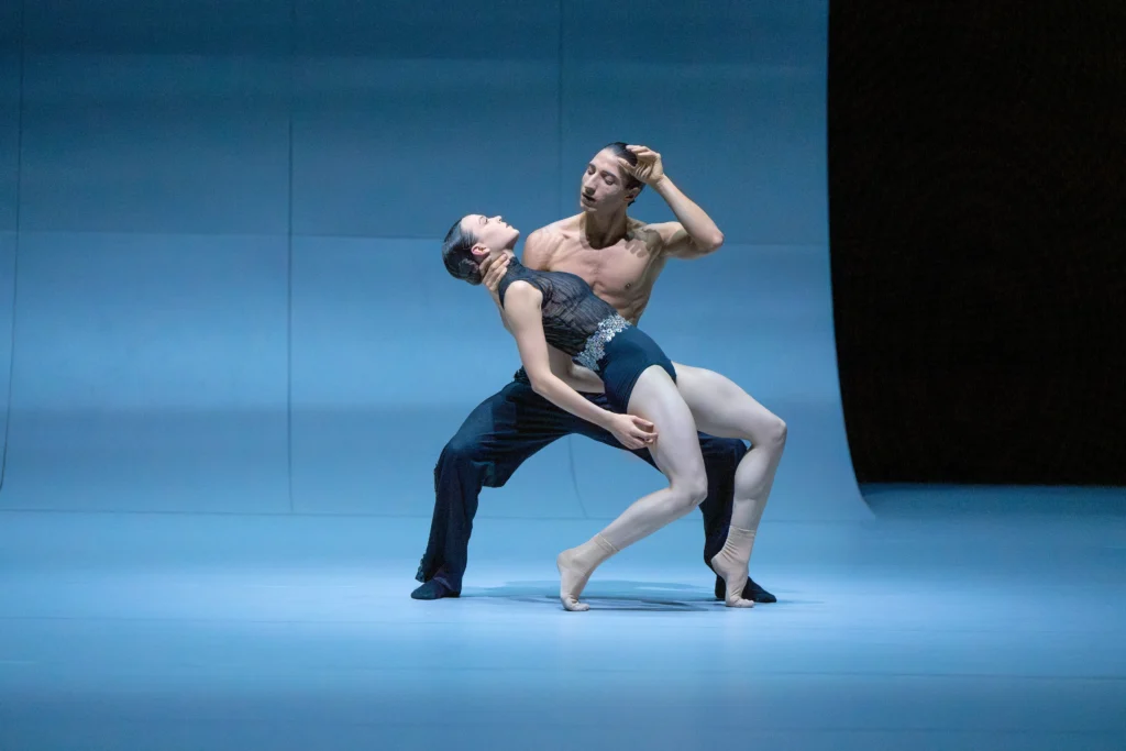 Mackenzie Brown, wearing socks and a patterned high-neck leotard in multiple hues of blue, perches on demi-pointe on bent legs and leans back as Martino Semenzato holds her by the back of the neck. He wears blue pants and squats in a low second position, looking towards Brown. They dance in front of a blue backdrop.