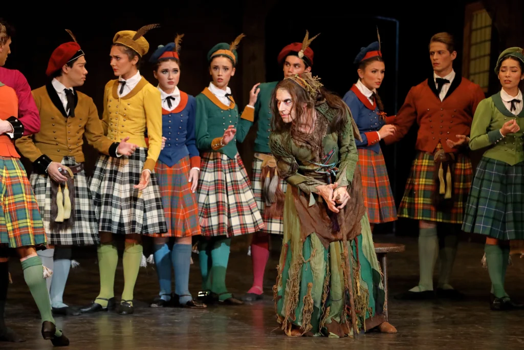 Ricki Bertoni, dressed as a witch in a tattered green and brown dress and long brown hair, stands onstage hunched over and holdign a cane. He looks devilishly towards stage right at someone off camera as a group of dancers dressed in multi-colored Scottish kilts, jackets and hats stand around him, looking concerned.