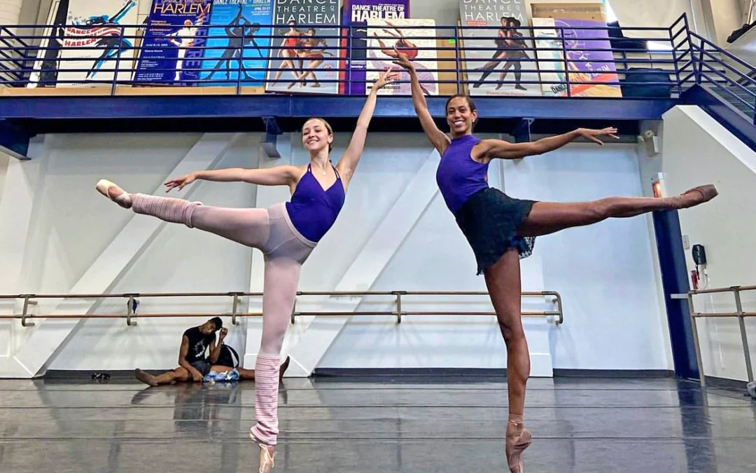 Alexandra Jones and Lindsey Donnell do a piqué first arabesque on pointe facing into each other in a large dance studio, with Jones on the left and Donnell on the right. Jones wears a royal blue leotard with pink tights pulled over it, pink legwarmers and pink pointe shoes. Donnell wears a royal blue high-neck leotard, a short black ballet skirt, and brown pointe shoes. They look out towards the camera and smile.