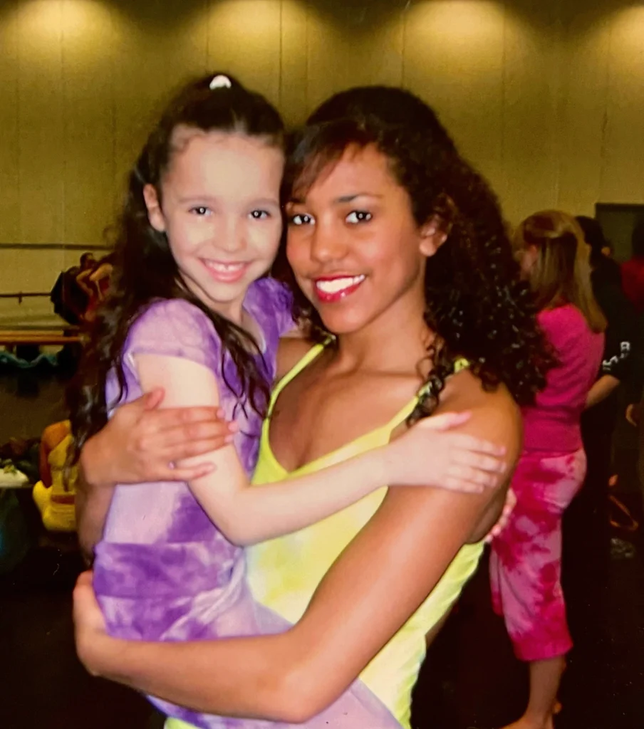 A young woman in a yellow leotard holds a femals child dressed in a purple tie-dyed leotard and leggings. They rest their heads against each other and smile.