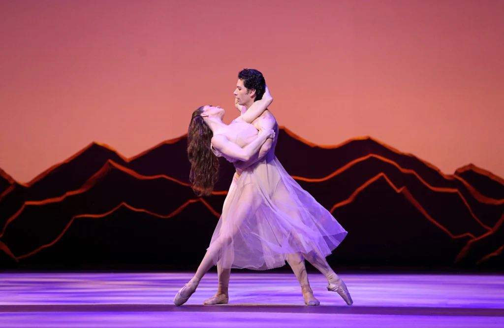 Cassandra Trenary and Herman COrnejo dance a passionate pas de deux onstage in front of a dark, jagged set. A shirtless Cornejo holds Trenary—who wears a light-colored, gauzy gown—closely around her waist as she wraps her left arm around his neck and grasps his left arm. She stretches her legs into a wide, parallel fourth, her feet barely touching the ground. Her long hair falls down her back as she leans back and looks up at him.
