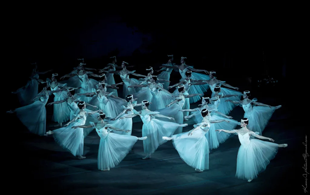The National Ballet of Ukraine's corps de ballet of women performs a romantic ballet onstage, in dark lighting. They wear sylph-like costumes.