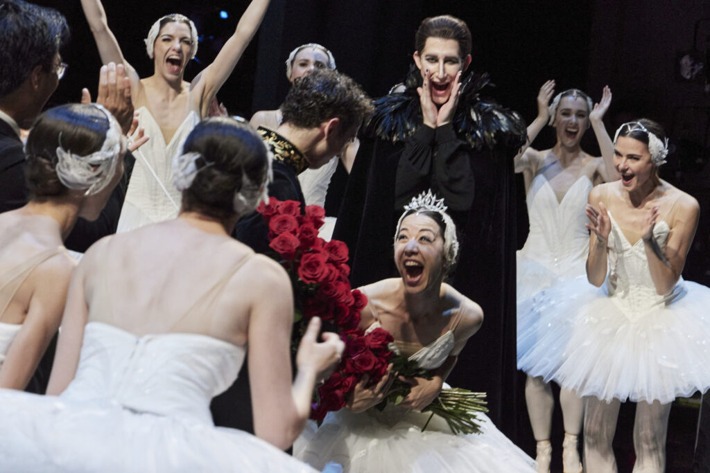 Jill Ōgai and Marcus Morelli hold roses as they celebrate their promotions onstage. Ōgai leans forward and opens her mouth in radiating joy, the other company members celebrating, cheering, and dancing around them both.