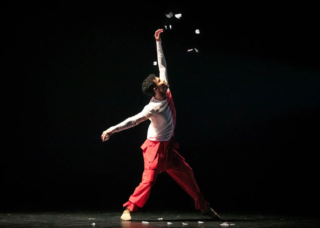 Fábio Mariano performs onstage. Small pieces of paper appear on the stage and above his left arm, which is stretched upward. He is wearing red pants and a red and white top.