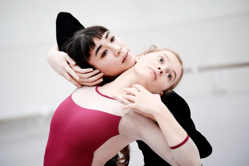 Brett Fukuda works with Lara Wolter in rehearsal. They are shown waist-up, and they lean on each other's shoulders facing backward from each other.