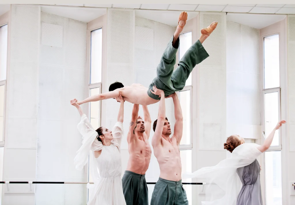 The main cast of "Muse Paradox" rehearses in a large, white studio. a woman and two men lift a third man above their heads, supporting his hips and shoulders, as a second woman dances off to the side.