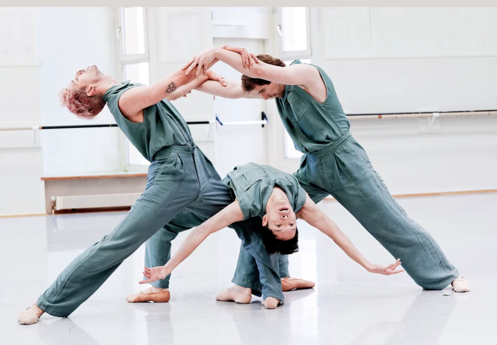 Three men in teal jumpsuits dance a pas de trois. Two stand and lunge toward each other, holding the others' arms, as the third bends backward, his back resting on their bent knees.