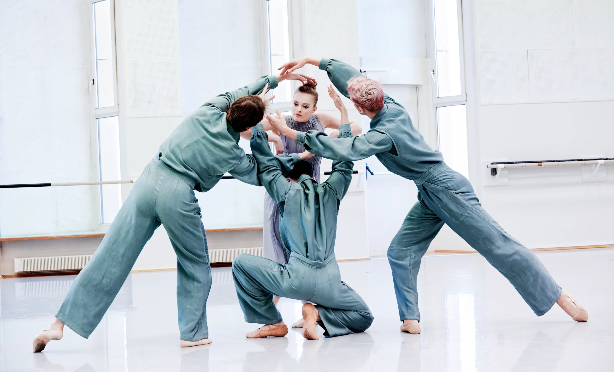 In a spacious, all-white dance studio, three male and one female dancer rehearse a new take on "Apollo" with the woman as the central figure. The men lean in toward her in crouches and lunges, and she peers through the negative space left by their arms, circling her head.