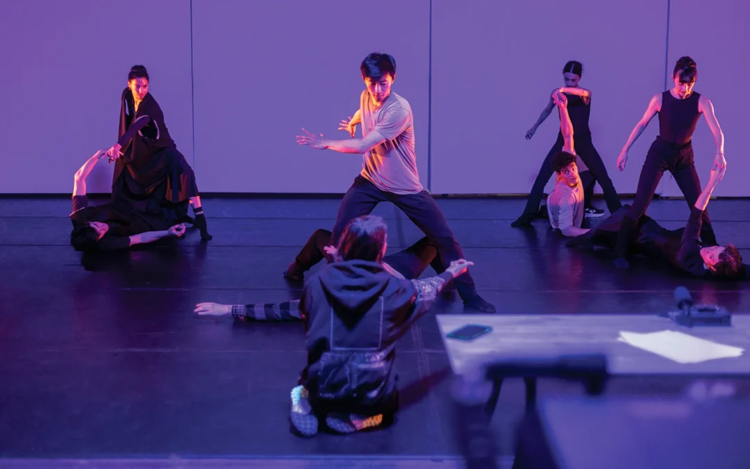 Aszure Barton, in a large sweatshirt, kneels in front of four couples as she leads a rehearsal in a dark studio lit by purple-colored gels.