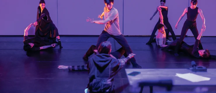 Aszure Barton, in a large sweatshirt, kneels in front of four couples as she leads a rehearsal in a dark studio lit by purple-colored gels.