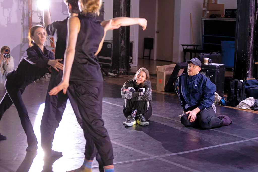 Aszure Barton and James Gregg sit on the floor while watching three dancers in rehearsal. Two women flank a man, the woman furthest from the camera lunging in toward him. In the background, a woman poses with a camera.