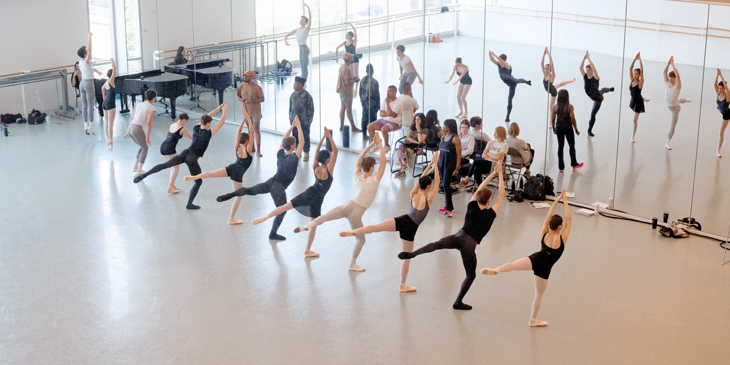 Twelve young male and female dancers are shown photographed from above and from a distance. They form a straight line at the front of the large dance studio, facing a wall of mirrors. The two dancers on the far stage left end of the line jump up, their legs in paralell and their arms at their sides, while the next two plié. The rest of the dancers pose in arabesque plié with their left leg up and their arms lifted above their heads, hands clasped. A team of adults face the dancers and watch them; some sit in chairs while the others stand. The female dancers wear leotards, short ballet skirts, pink tights and pointe shoes, while the male students wear t-shirts, black or gray tights and ballet slippers.