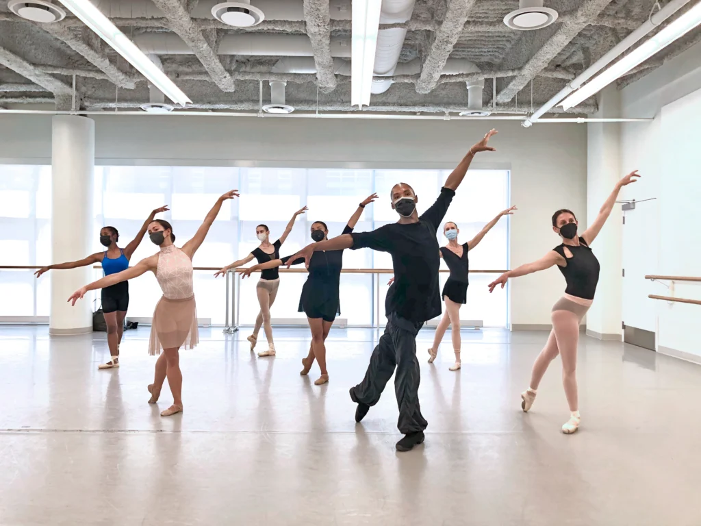 Six adult ballet students stand in staggered lines behind their teacher in a spacious dance studio. They all pose in tendu derriere croisé with their left leg back. They lift their left arm in a high diagonal and open their right arm directly to the side, and look out over their right shoulder. The dancers and teacher wear various dance clothing and face masks.