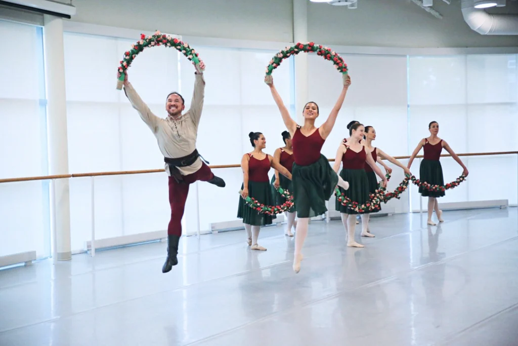 A male and female adult ballet student do a jubilant arabesque temps levé while holding a flower garland above their heads. The man wears a khaki-colored long-sleeved shirt, burgundy tights and black ballet boots, while the woman wears a burgundy leotard, pink tights and a knee-length black ballet skirt. Behind them, six women in the same dancewear pose in B plus with their right foot behind them, holding their arms in demi-second with the garland in their hands.