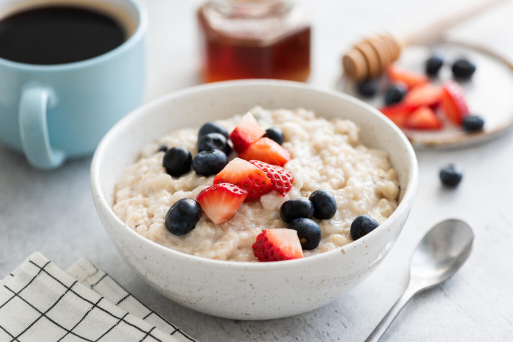 Oatmeal porridge with berries, honey and cup of coffee on white table. Healthy breakfast food, vegetarian porridge oats bowl