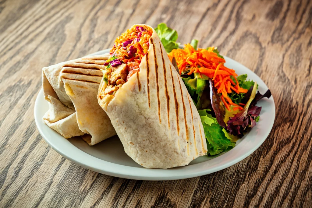 A grilled vegetable and chicken wrap with a side salad rests on a white plate on top of a wooden table top.