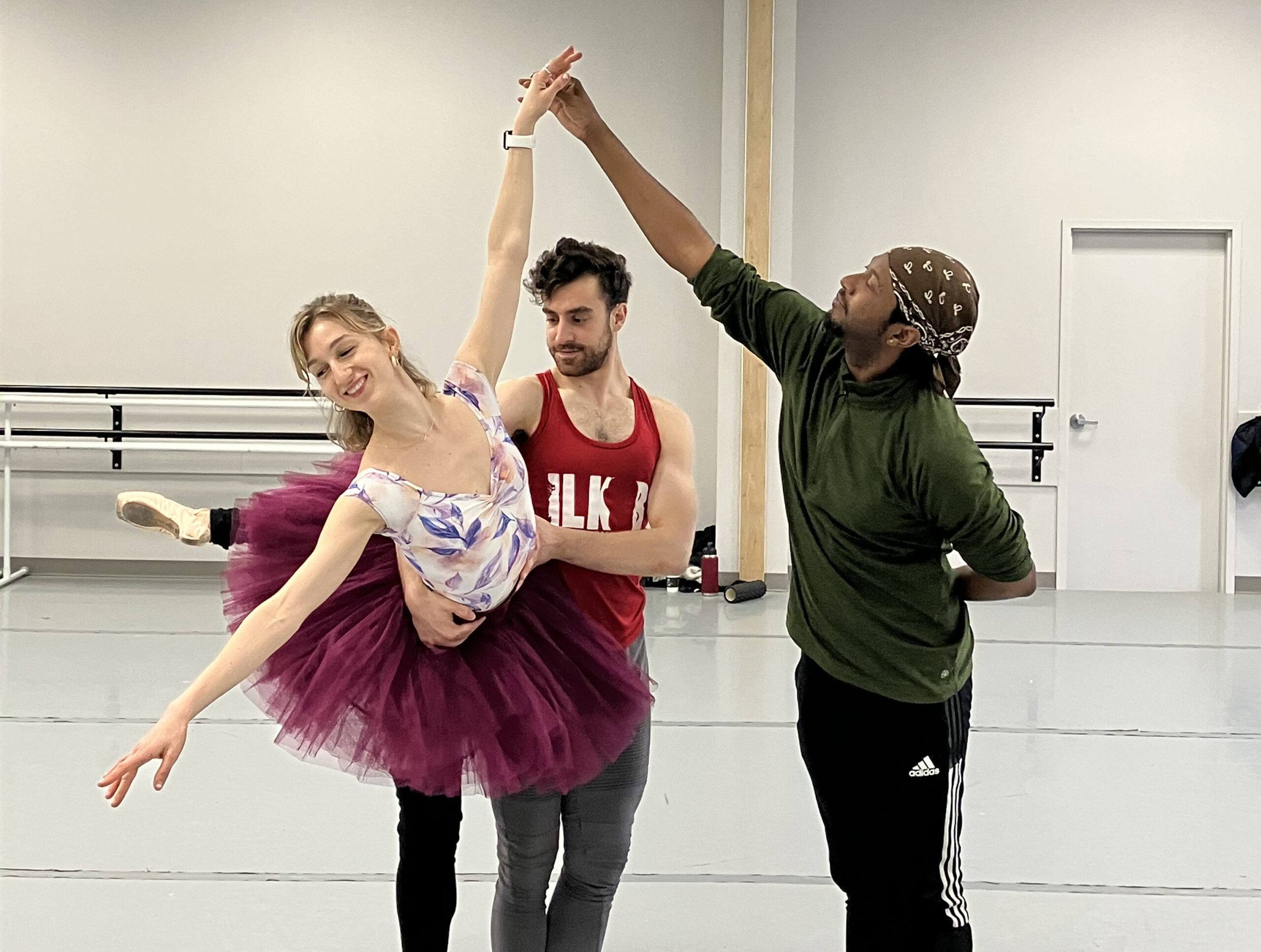 Lucy Hamilton and Lukas Figliozzi practice a partnering move while Jayson Douglas reaches up and adjusts Hamilton's left hand. Hamilton poses in attitude derriere croisé with her left leg lifted as Figgliozzi stands on her left side and holds her around the waist with both hands. She wears a burgundy practice tutu, black legwarmers, pointe shoes and a white leotard with purple flowers. Figliozzi wears a red sleeveless T-shirt and gray tights, while Douglas wears a green long-sleeved shirt, black athletic pants, and a brown bandana around his head.