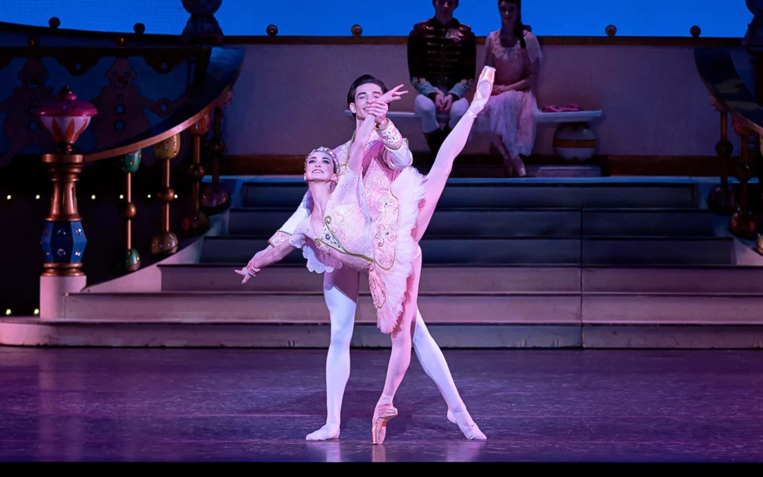 Jessica McCann, wearing a pink tutu, pick tights and pointe shoes, does a penché on pointe, looking out towards the audience. He raises her left arm high and her right arm low while her partner, Colin McCaslin stands behind her and holds both of her wrists for balance. He wears white tights and ballet slippers and a light-colored jacket with gold-trimmed cuffs.