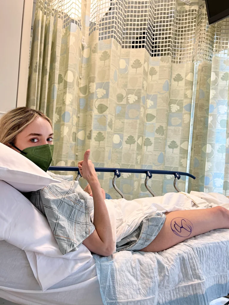 Jessica McCann lays on a hospital bed and turns her head towards her right to look directly at the camera, giving a thumb's up sign with her right hand. She wears green face mask and a hospital gown that is drawn up high to show her right leg. Her right outer thigh has a circle drawn in black marker. She is surrounded by a hospital curtain with a green, blue and light brown leaf design.