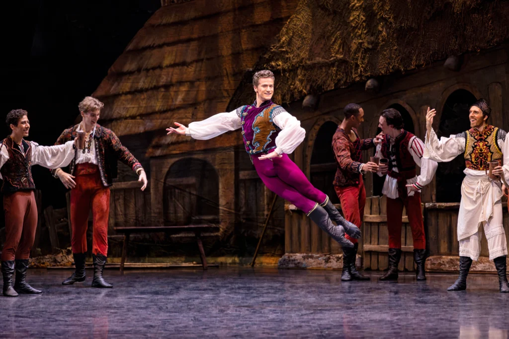 Jordan Viet performs a cabriole derriere during a ballet performance, with his left hand on his hip and his right arm out to the side. He looks out toward the audience and smiles confidently. He wears a white peasant blouse, a brown and magenta vest, magenta tights, and black ballet boots. He performs in front of a large set depicting a wooden house and fence. A group of male dancers in variuos costumes mill about behind him, having a good time.