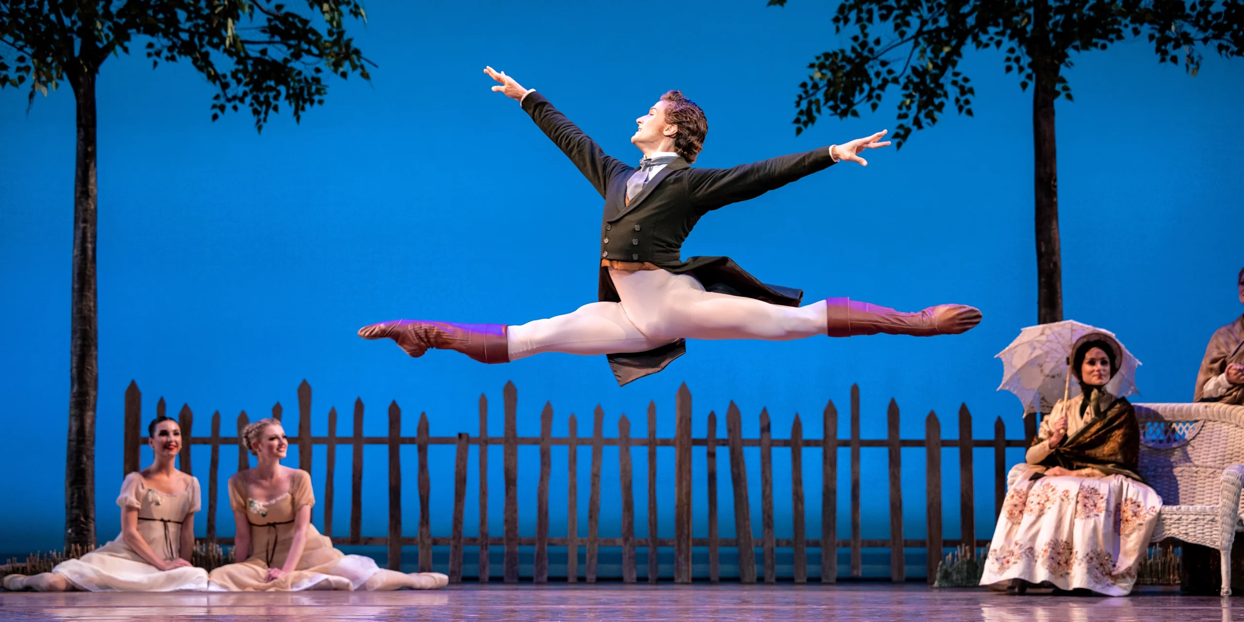 Jordan Veit leaps across the stage towards stage right during a performance of Onegin, his arms in first position. He wears a gray tie, dark sport jacket with tails, white tights, and brown ballet boots. He dances in front of a set that shows a picket fence and two trees in front of a blue sky. Two women in long white dresses sit on the ground upstage and watch Veit, while another sits on a wicker loveseat upstage left, holding a parasol and wearing a long dress, a shawl around her shoulders, and a bonnet.