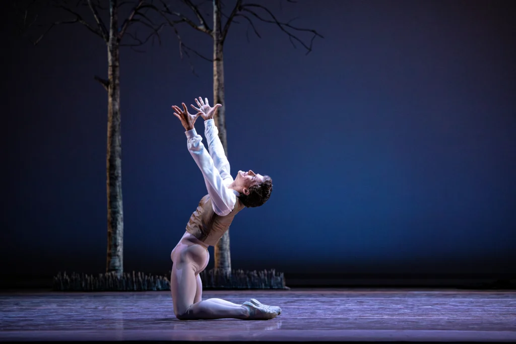 Jordan Veit is shown onstage in profile, on both knees and facing stage right. He reaches his arms skyward, his face in anguish. He wears a white blouse, tan vest, tan tights and ballet slippers. Upstage are two bare trees.