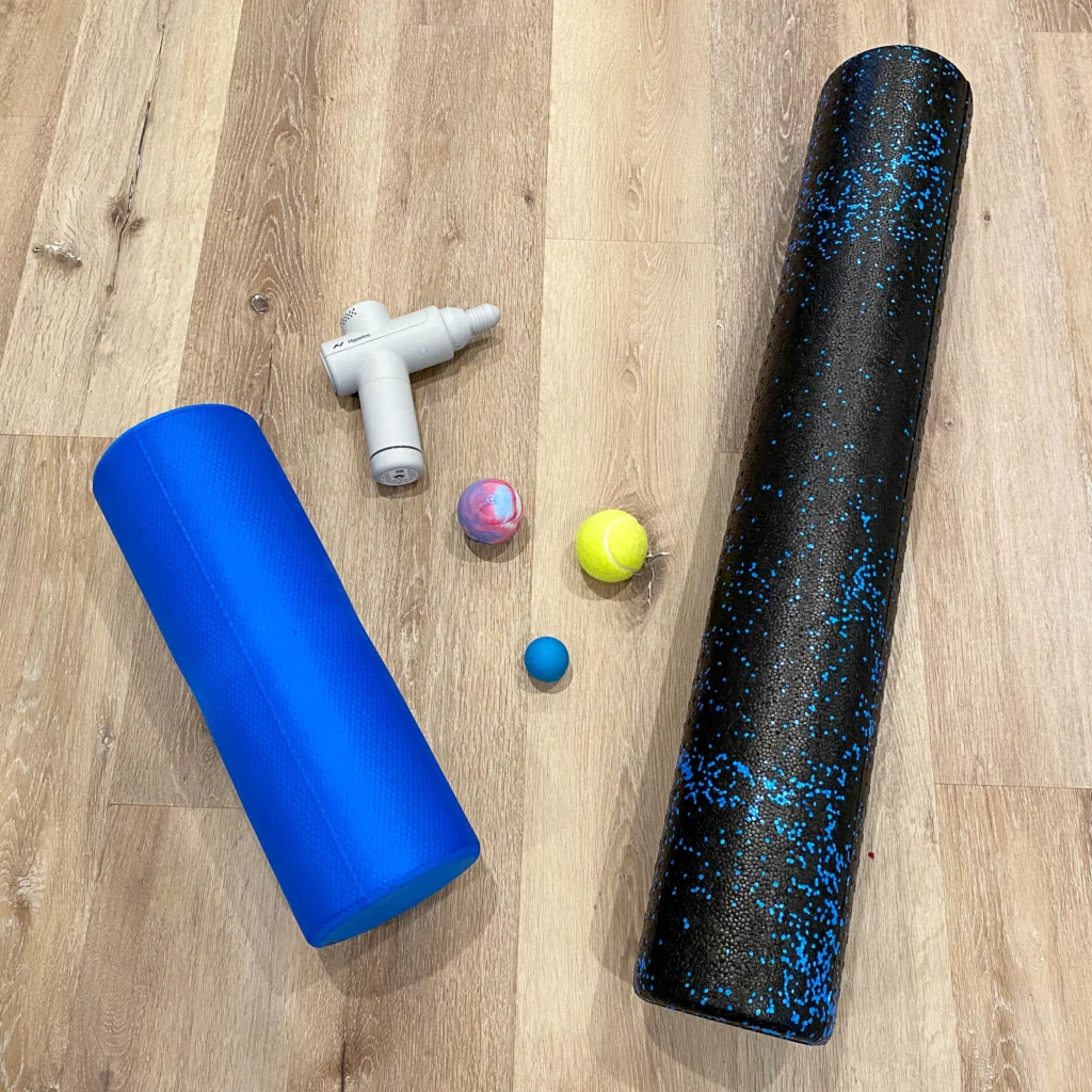A group of massage tools is shown from above on a wooden floor. They include two foam rollers, a tennis ball, two rubber balls, and a small white massage gun.