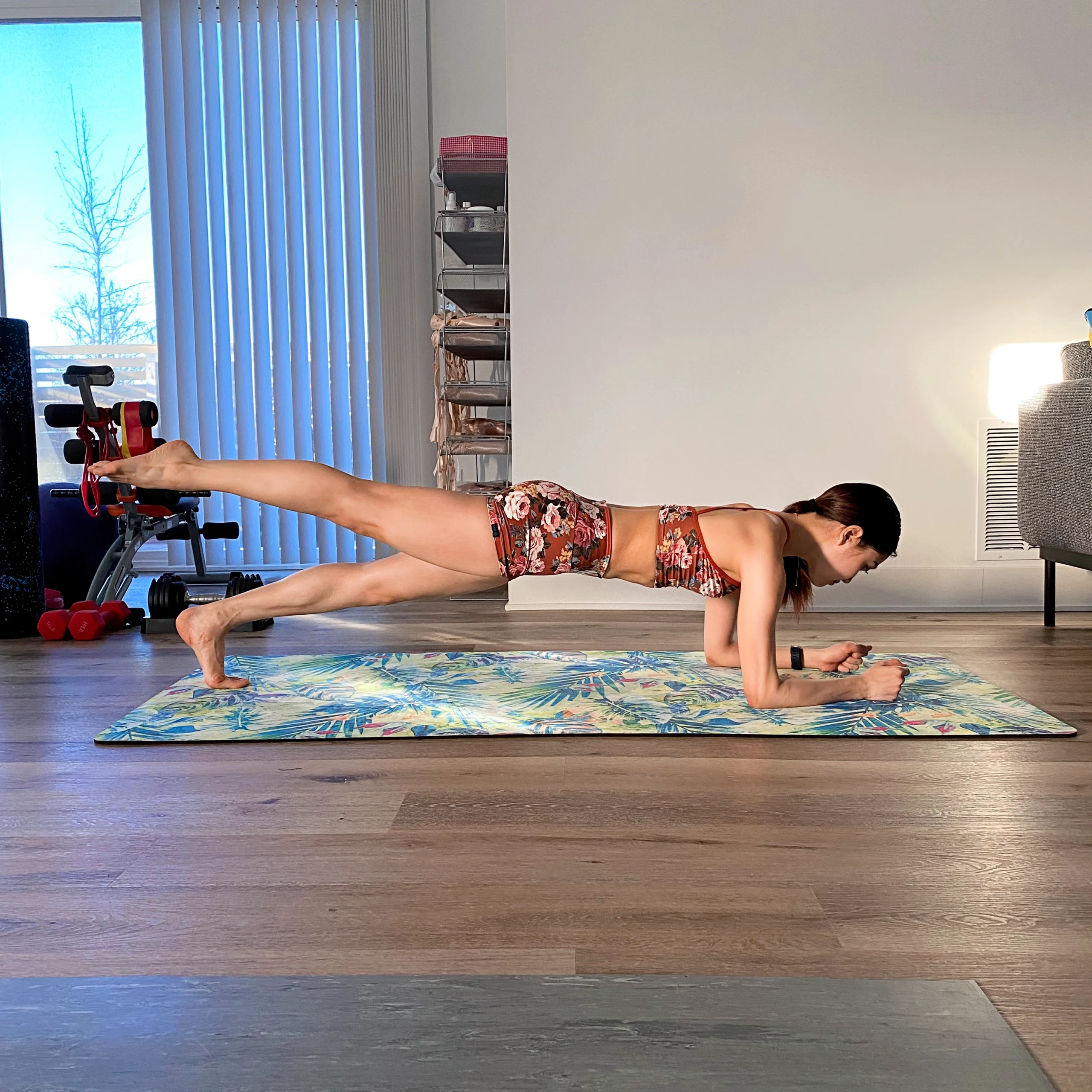 Yoshiko Kamikusa practices a plank exercise on a colorful yoga mat in her living room. She wears a pink and white floral sports bar and matching bootie shorts.