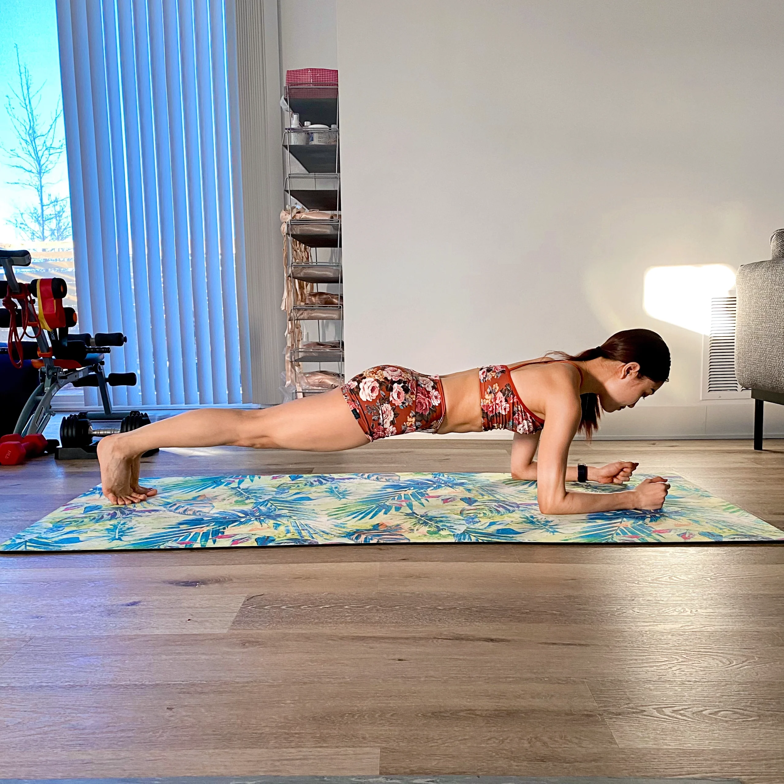 Yoshiko Kamikusa practices a plank exercise on a colorful yoga mat in her living room. She wears a pink and white floral sports bar and matching bootie shorts.