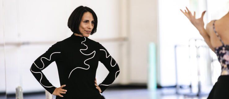 Annabelle Lopez Ochoa, wearing a black turtleneck with a squiggly white design on the front and the sleeves, is shown from the waist up with her hands on her hips. She is standing with her back facing the mirror in a ballet studio and watches dancers rehearse. On the far right of the photo, a female dancer's shoulder and hand are visible.