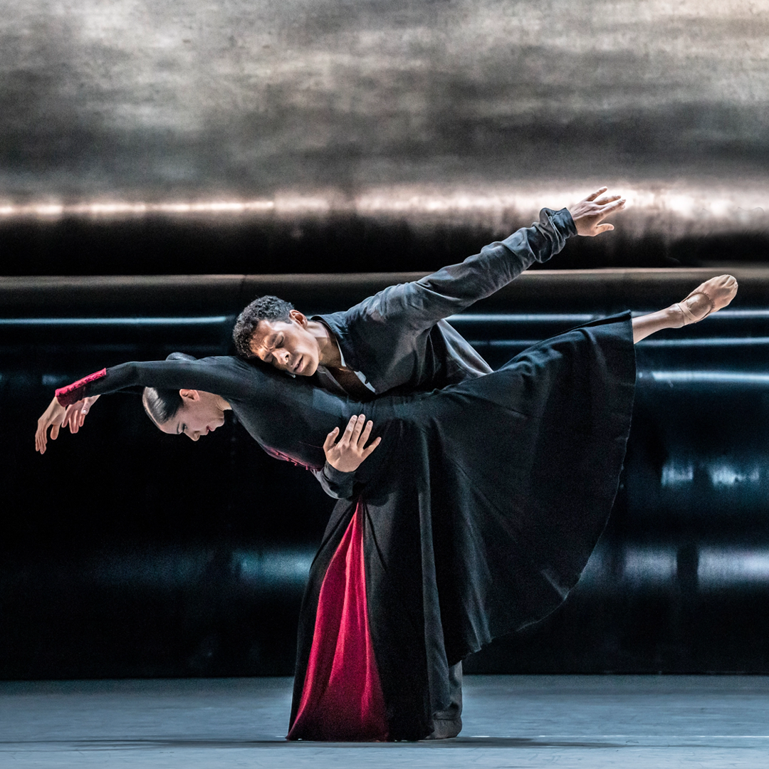 Celine Gittens and Tyrone Singleton perform a pas de deux onstage in front of a black and gray backdrop. Gittens des a deep arabesque elongé, bending forward at the hips and curving her arms above her head as Singleton holds her around the waist and tenderly leans his head against her mid-back. He wears a dark jacket while Gitttens is costumed in a long black dress with long sleeves and a dark red panel in the middle, and tan ballet slippers.