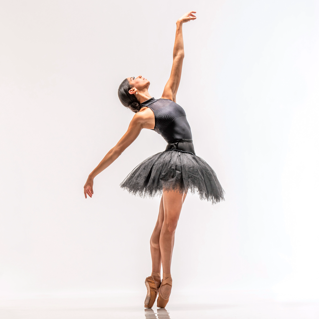 Céline Gittens, wearing a black high-neck leotard, black practice tutu and light brown pointe shoes, poses against a bright white backdrop. She stands on pointe in profile facing her left with her legs in sus-sous. She stretches her left arm up as she trailes her right arm low and slightly behind her. She bends slightly back, looking up towards the ceiling.