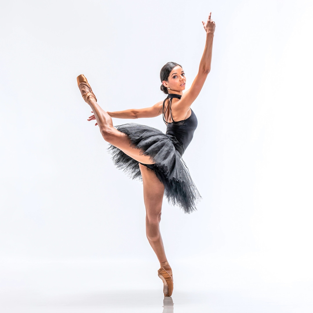Céline Gittens, wearing a black high-neck leotard, black practice tutu and light brown pointe shoes, poses against a bright white backdrop. She does a high attitude derriere in effacee with her right leg raised and her left foot on pointe. She lifts her curved right arm up and slightly in front of her face, and reaches her left arm back and to the side. She wears her hair in a low chignon and looks out at the camera.