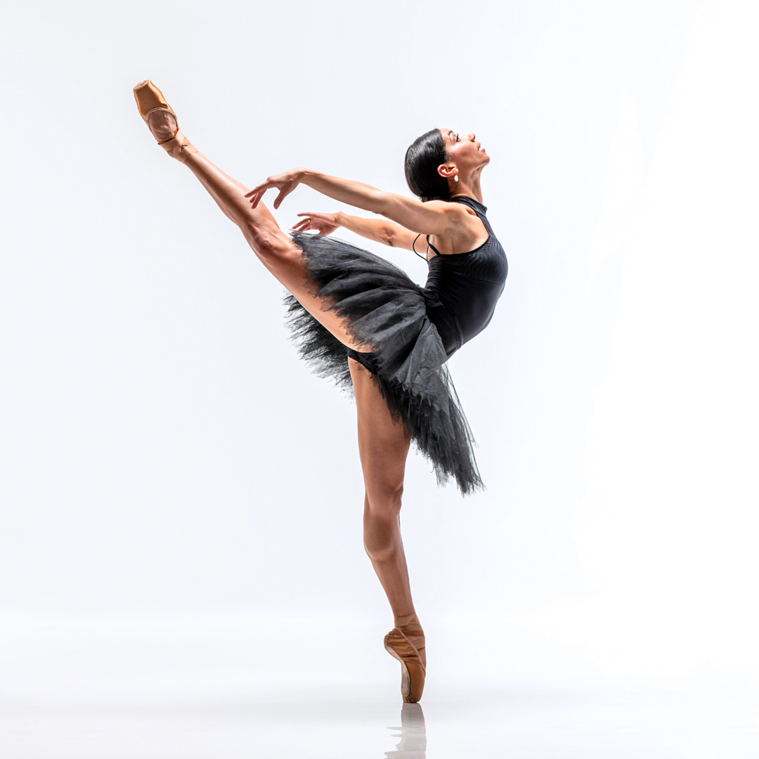 Céline Gittens, wearing a black high-neck leotard, black practice tutu and light brown pointe shoes, poses against a bright white backdrop. She poses in profile facing her left in an attitude on pointe with her right leg raised. She presses both arms back and arches her upper body, looking up towards the ceiling.