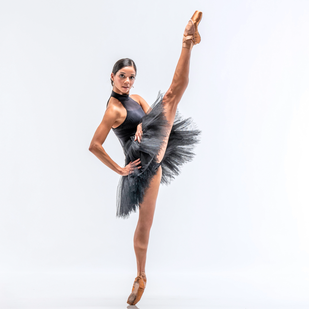Céline Gittens, wearing a black high-neck leotard, black practice tutu and light brown pointe shoes, poses against a bright white backdrop. She stand on pointe in profile facing her left side and does a grand battement front with her right leg. She puts her right hand on her hip and rests her left hand on top of her tutu, and looks directly at the camera with a small smile.