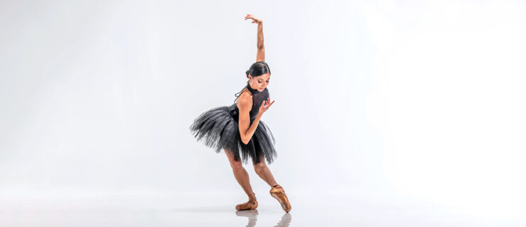 Céline Gittens, wearing a black high-neck leotard, black practice tutu and light brown pointe shoes, poses against a bright white backdrop. She does a tendu devant in effacé with her left leg, bending her right knee in plié, and reaches her upper body forward as she looks down at her pointed left foot. She brings her right amr in towards her chest and reaches her left arm straight up.