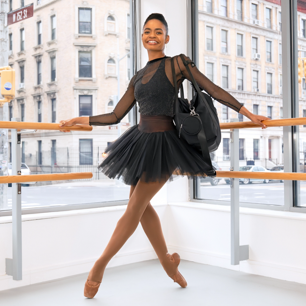Alexandra Hutchinson poses in the corner between the ends of the barres in a Dance Theatre of Harlem studio. She is wearing a sparkling sheer black top over a black leotard, with a black tutu and black dance bag. She is smiling at the camera.
