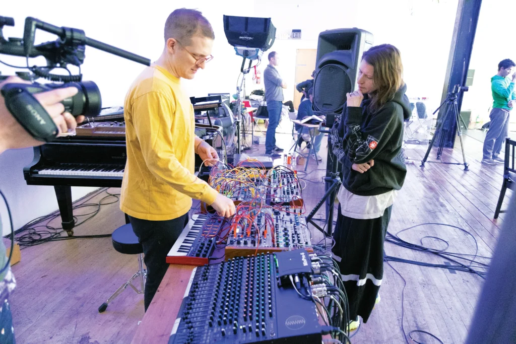 Sam Shepherd and Aszure Barton stand on either side of a large soundboard setup. Shepherd looks down at his fingers on a button, and Barton looks at it pensively.