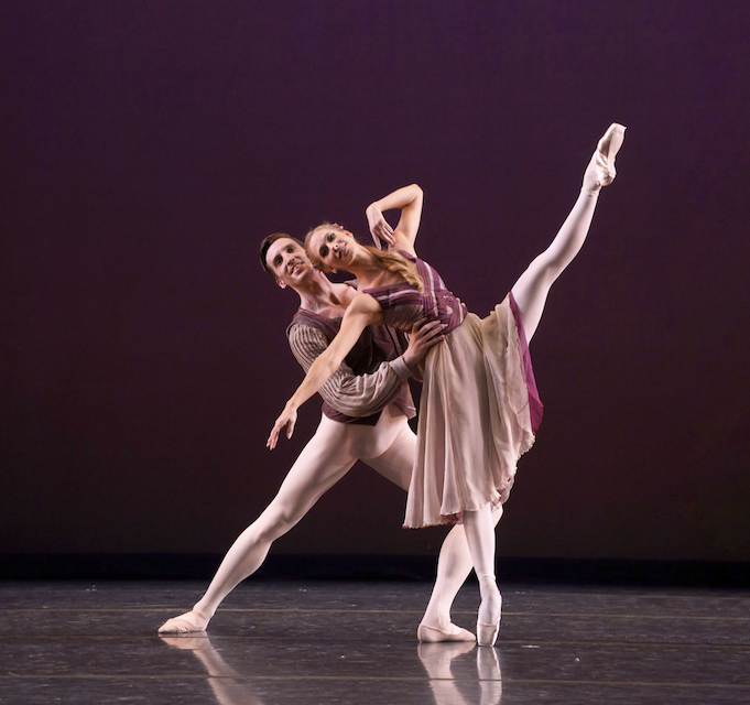 Danielle Brown and Richard House perform a pas de deux onstage in front of a plum-colored backdrop. Brown fondus and extends her left leg upward, leaning back as House supports her waist and poses in a lunge.