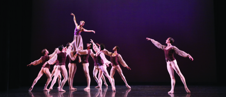 Onstage, an ensemble of dancers perform a ballet in front of a plum-colored backdrop. They wear purple tunics and white tights, or purple flowing dresses.