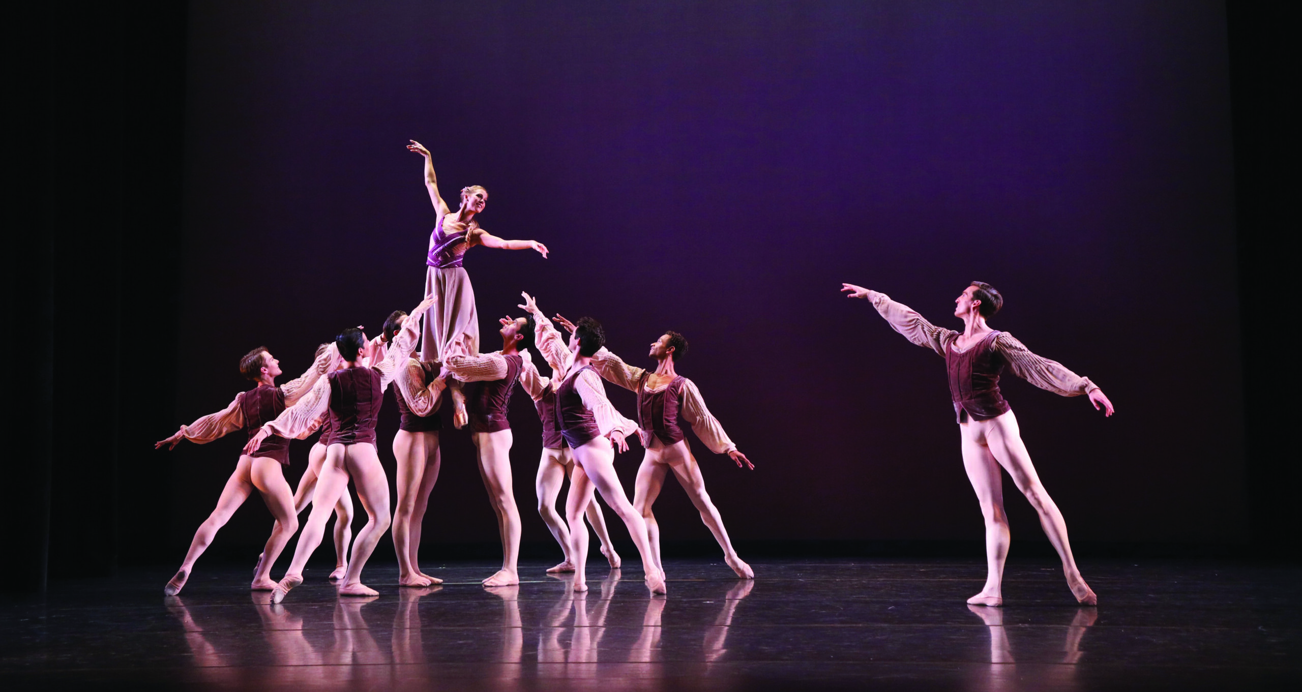 Onstage, an ensemble of dancers perform a ballet in front of a plum-colored backdrop. They wear purple tunics and white tights, or purple flowing dresses.