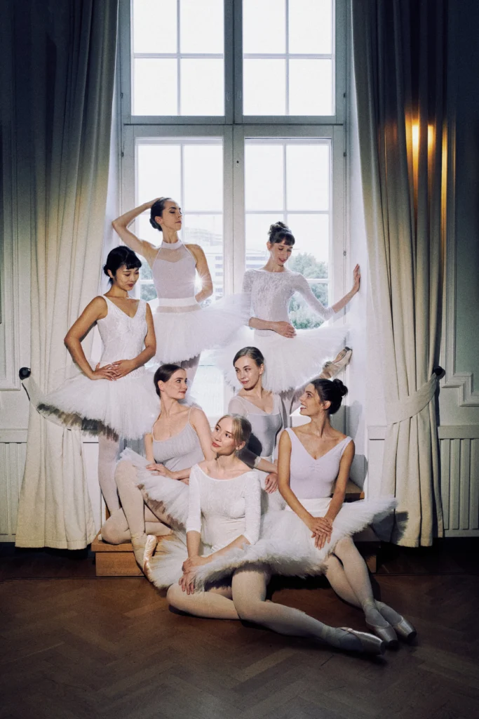 A group if seven ballerinas in white tutus, pink tights and pointe shoes make a close formation in front of a tall window with long white curtains on each side. Three dancers stand close to the windo pane, while the other four sit at various levels and in different poses. They all look at each other and smile.