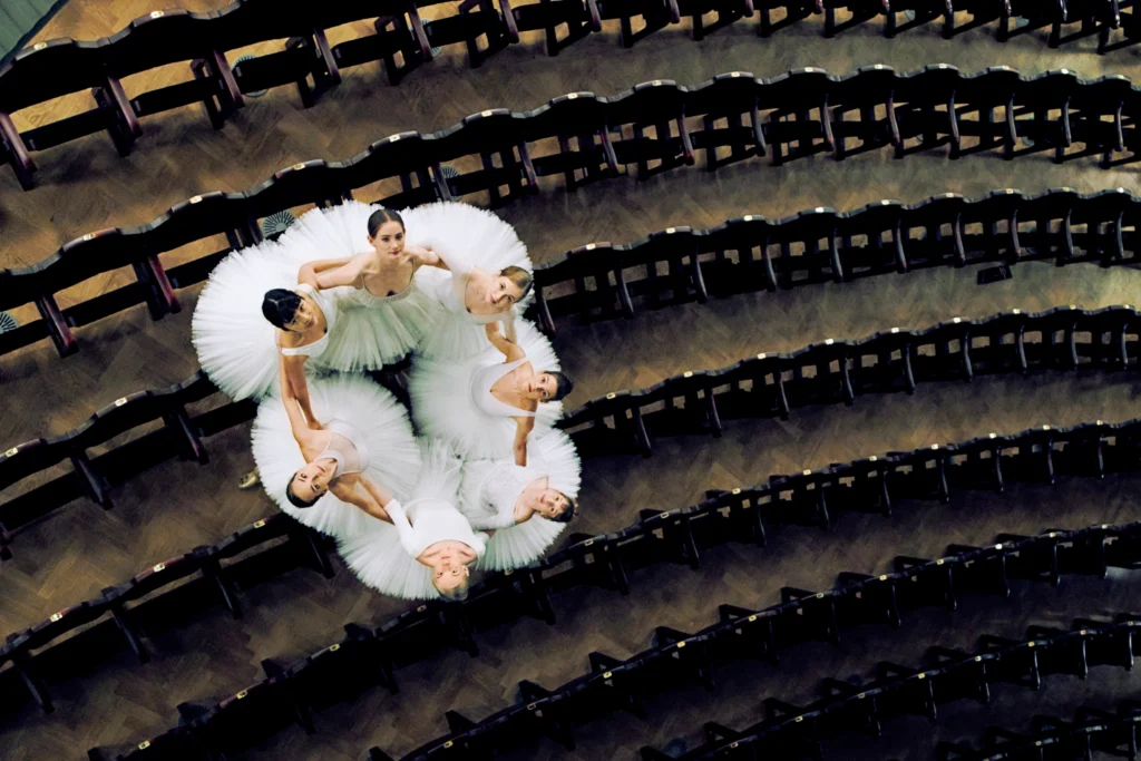 A group of seven ballerinas in white tutus is shot from above in the house of a theater. They wrap their arms around each others' waists and form a circle among the rows of seats. They look up and smile towards the camera.