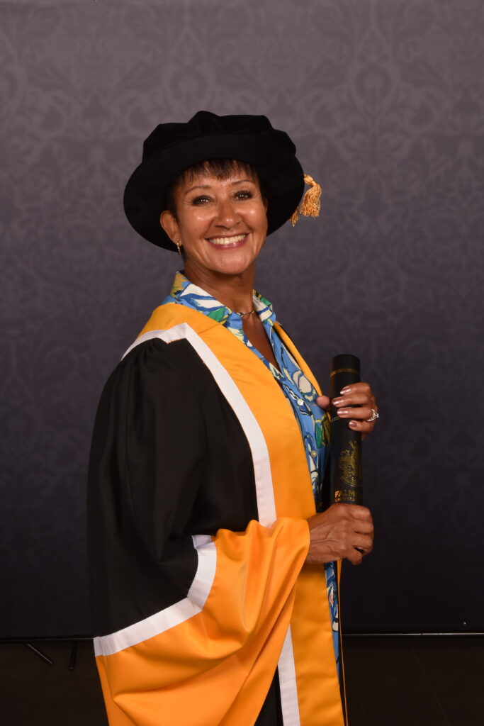 Julie Felix, shown wearing decorous master's graduation attire, smiles and poses for a photo while holding her honorary diploma.