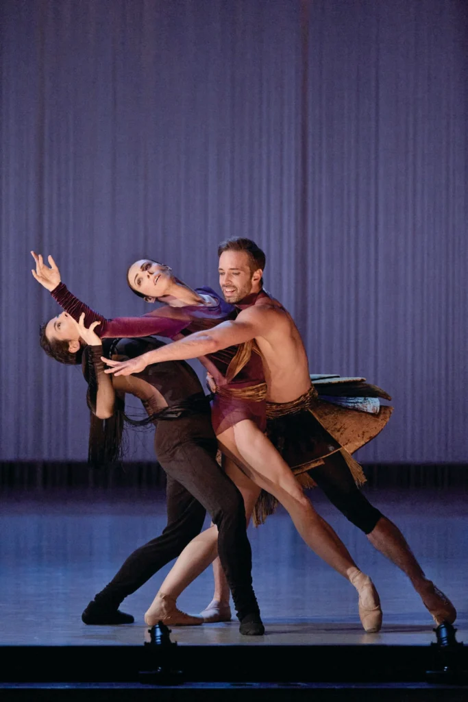 A pas de trois of two men and a woman perform onstage, leaning into each other and reaching up to the sky or across each others' bodies. The woman, in the middle, and the man to her right both wear tutus. The man on the far left wears a black unitard.