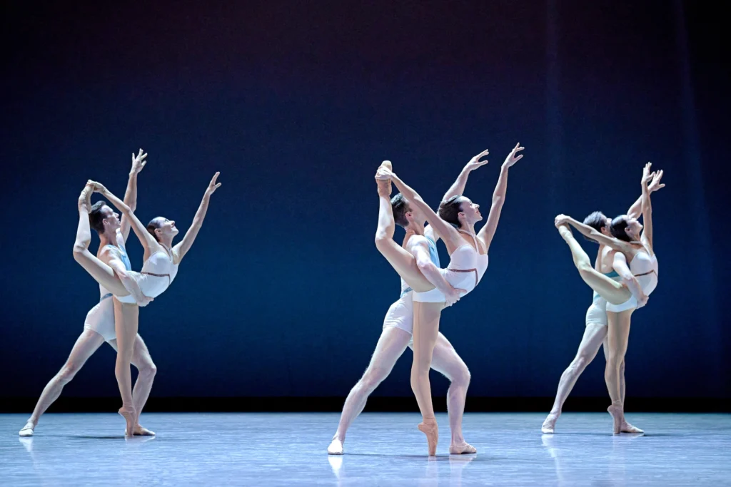 Three pairs of male and female couples stand in a triangle formation, the women standing on pointe and holding their back leg in a high attitude; the men support their backs and reach up with their arms to match the line.