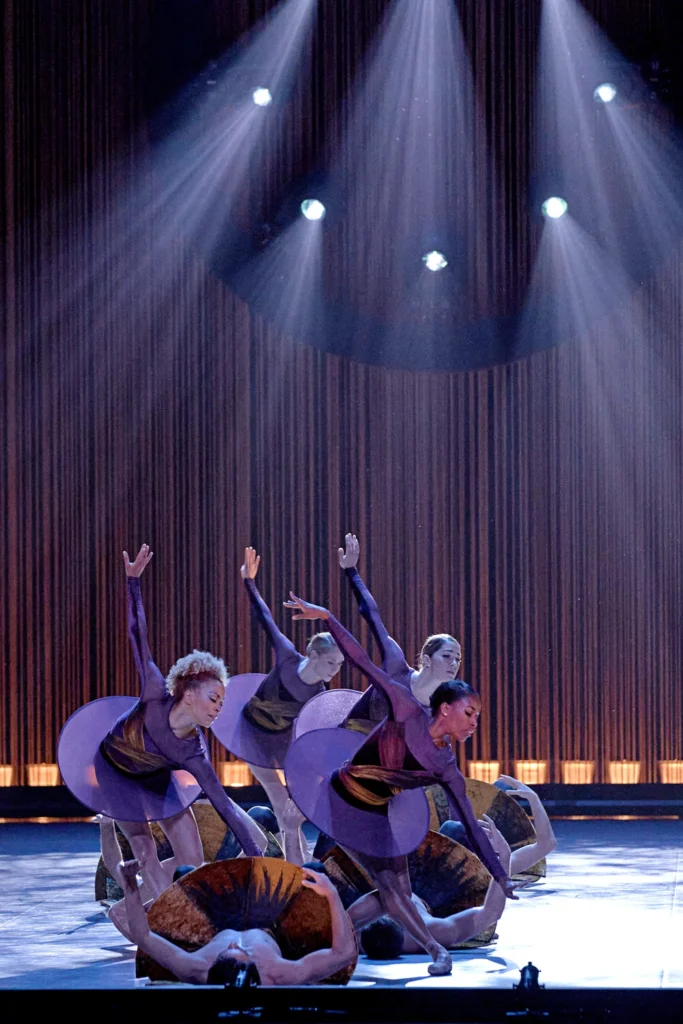 A group of four women in dark maroon tutus stand in a fondu tendu croise as they reach down toward four men laying on the floor below them, backs arched and hips bridged up.