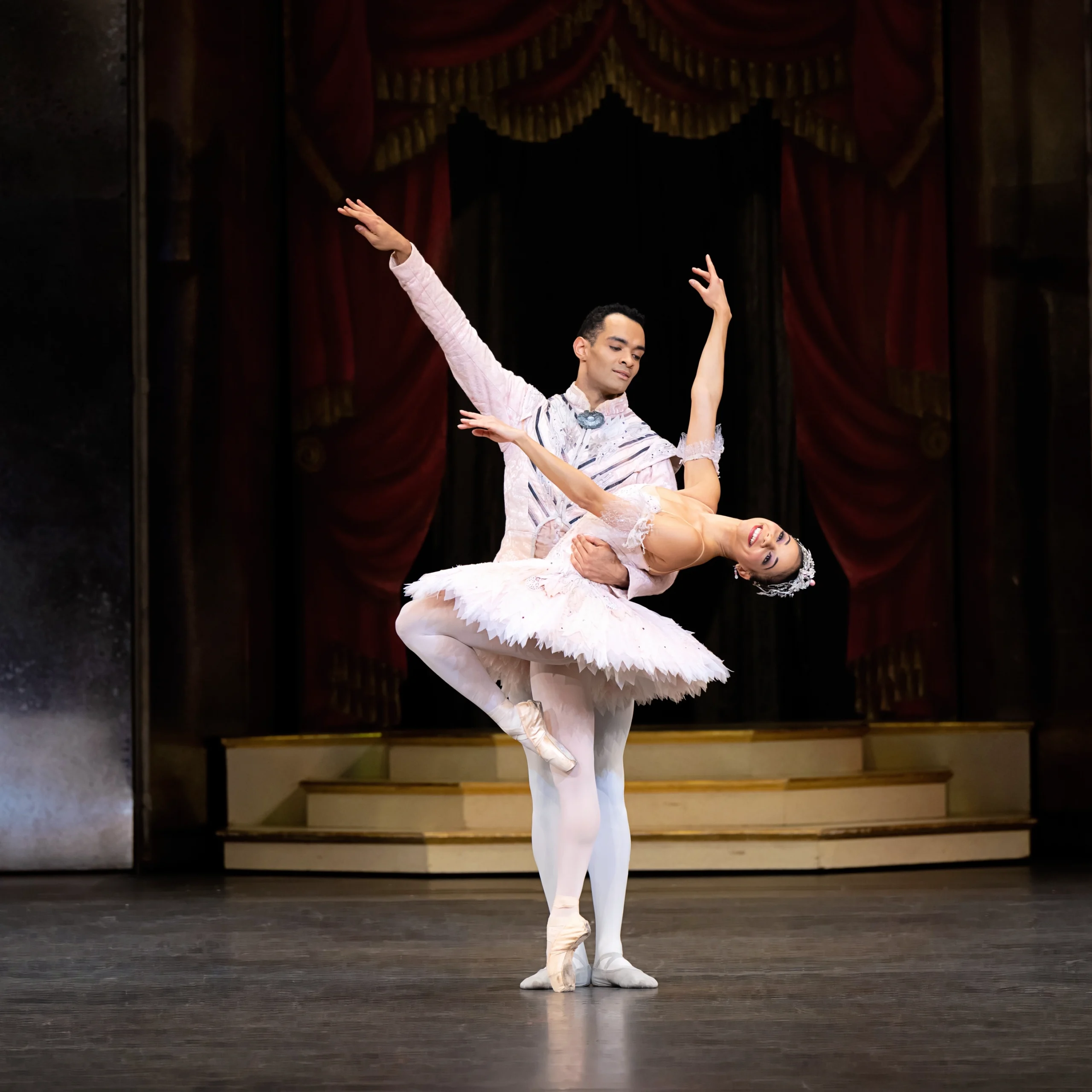 Céline Gittens and Brandon Lawrence perform a pas de deux during a performance of the Nutcracker. Gittens stands on pointe facing stage right with her left leg in a parallel passé, leaning back deeply as Lawrence stands behind her and holds her waist. Gittens wears a light pink tutu, pink tights and pointe shoes and a silvery crown and holds her arms in third position, looking out to the audience with a broad smile. Lawrence, in a light pink and silver tunic, white tights and white ballet slippers, looks down at her affectionately and lifts his right arm up.