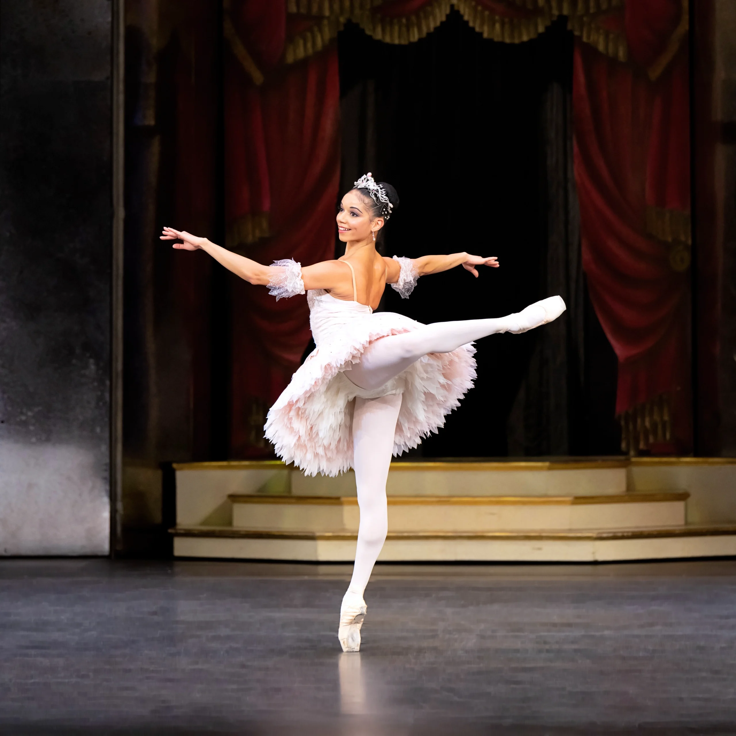Céline Gittens dances a solo during a performance of Nutcracker. She faces the upstage right corner and poses in attitude back, her arms in second position as she looks back over her left shoulder. She wears a light pink tutu, pink tights, and pink pointe shoes, and a silvery tiara. She has a bright smile on her face.