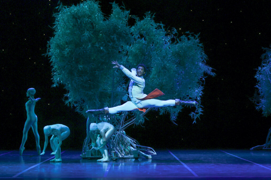 GIan Carlo Perez leaps onstage in front of a lush tree. He carries his arms in second arabesque position and looks out towards the audience. He wears a white jacket with long tails, white tights and black boots. Two female dancers in green unitards and skulls caps stand near the tree and bend their knees, crouching slightly, while another further upstage poses in fourth position on pointe, her amrs out to the side.